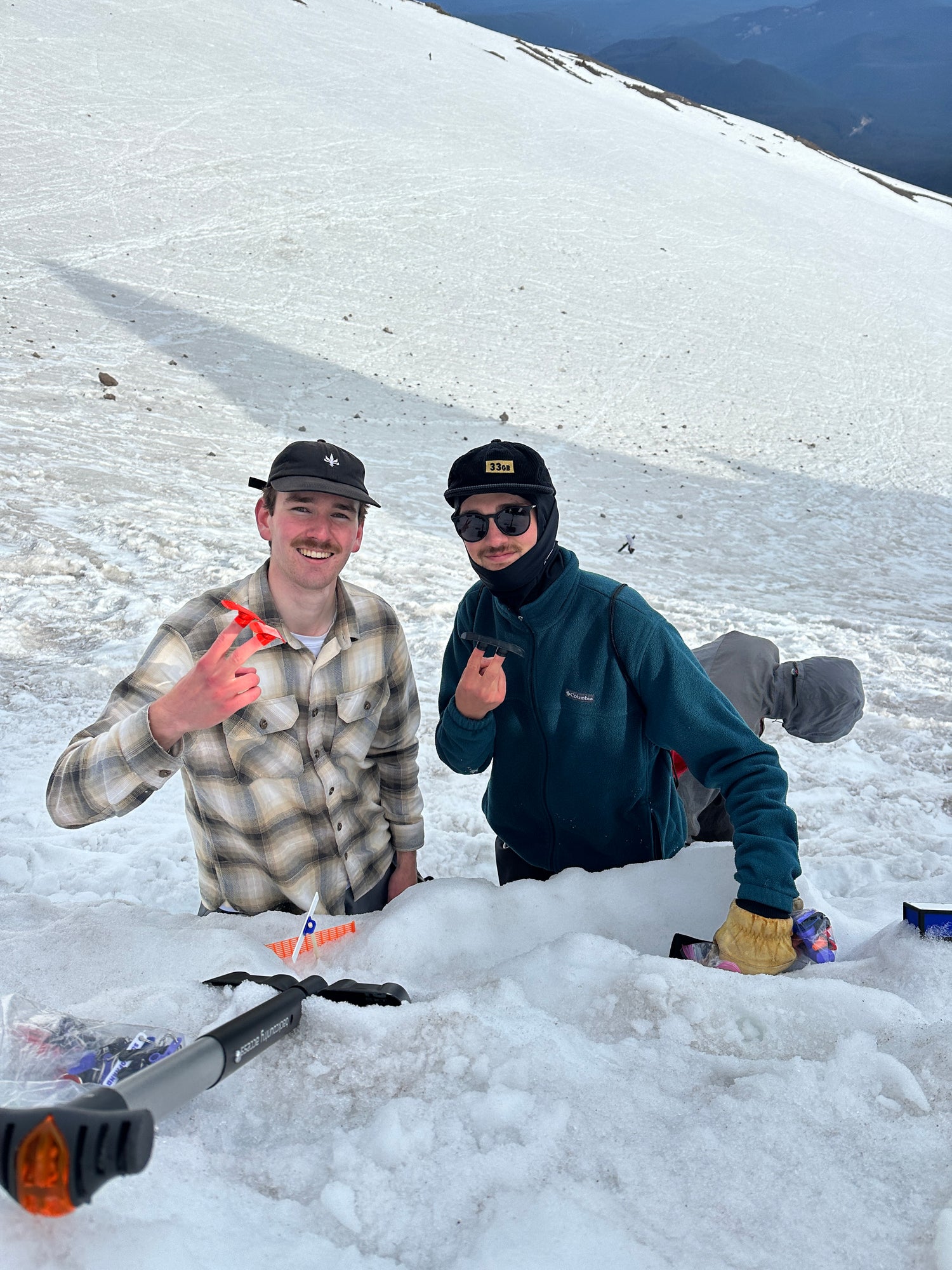 Evan (left) and Austn (Right) holding up peace signs with a pair of finger skis and a snowboard at the Mt. Hood Summer Solstice Party at Illumination Rock 2025.
